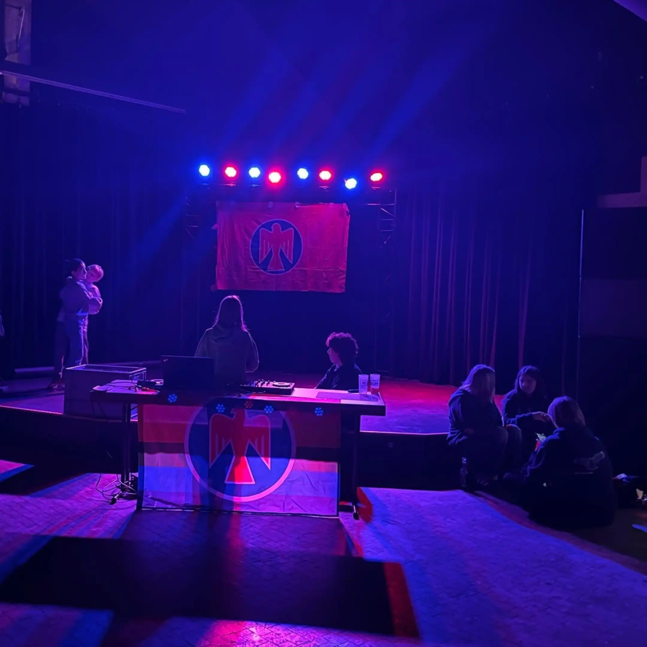 Im Vordergrund steht ein Tisch mit Falkenfahne in Regenbogenfarben geschmückt. Auf diesem stehen Laptop und DJ-Console. Dahinter stehen zwei Personen. Hinter dem DJ-Pult ist die Bühne auf der ein Paar tanzt. Rechts der Bühne sitzt eine Gruppe von drei Personen am Boden
