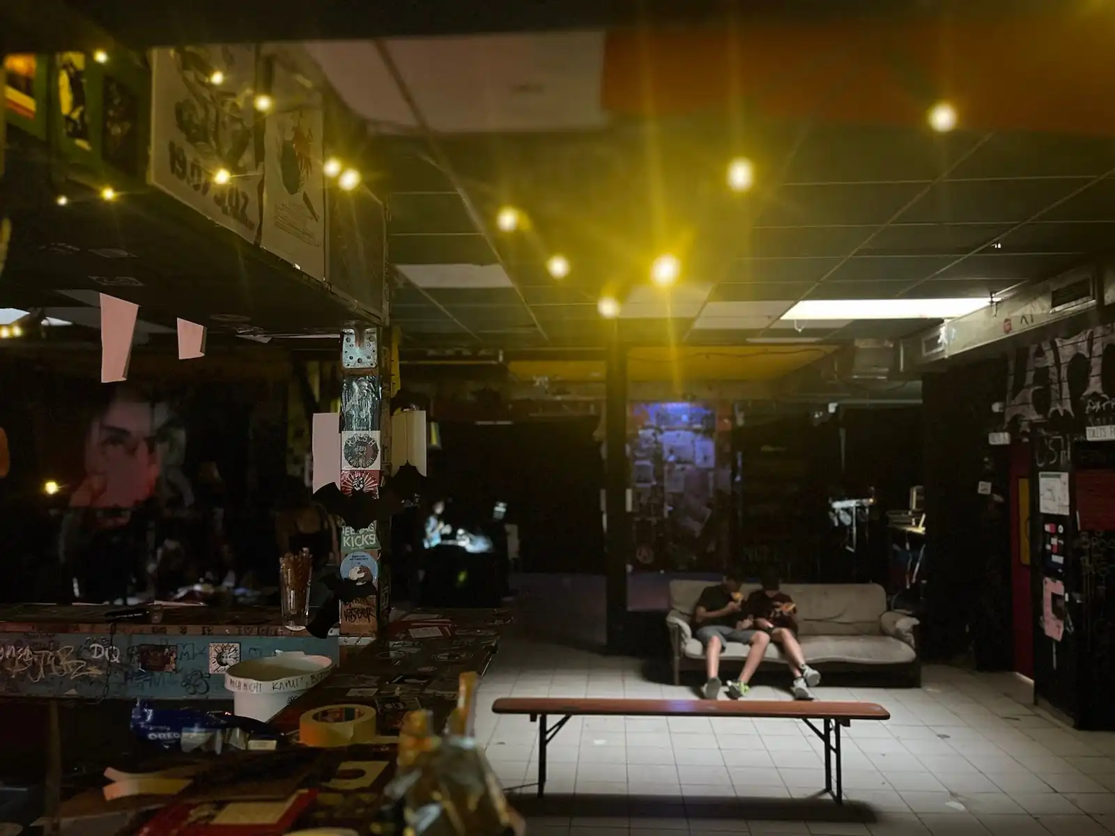 Blick von einer vollgestellten Bar auf einen gefließten Partyraum mit einer Bierbank in der Mitte. Auf einer Couch im Hintergrund sitzen zwei Jugendliche. Im oberen Bildrittel hängt eine Lichterkette.