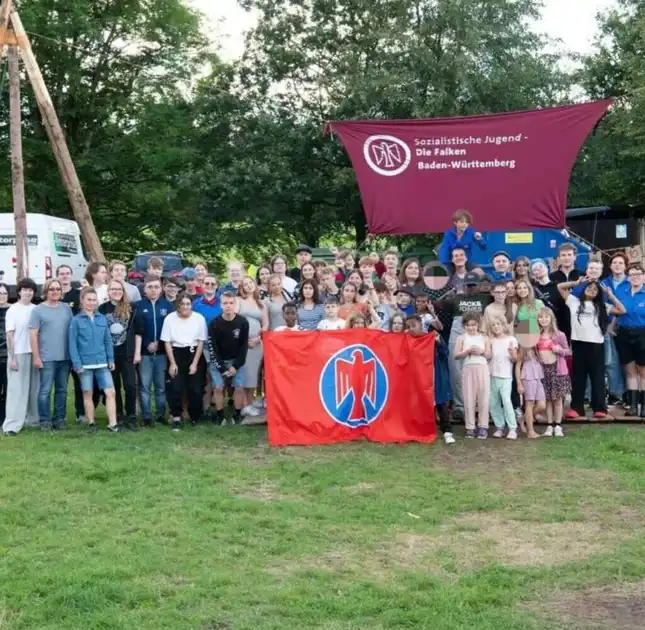 Eine Gruppe von Kindern und Erwachsenen steht mit Falkenfahnen auf einer Wiese. Über ihren Köpfen ist ein rotes Banner gespannt mit der Aufschrift SJD-Die Falken Baden-Württemberg