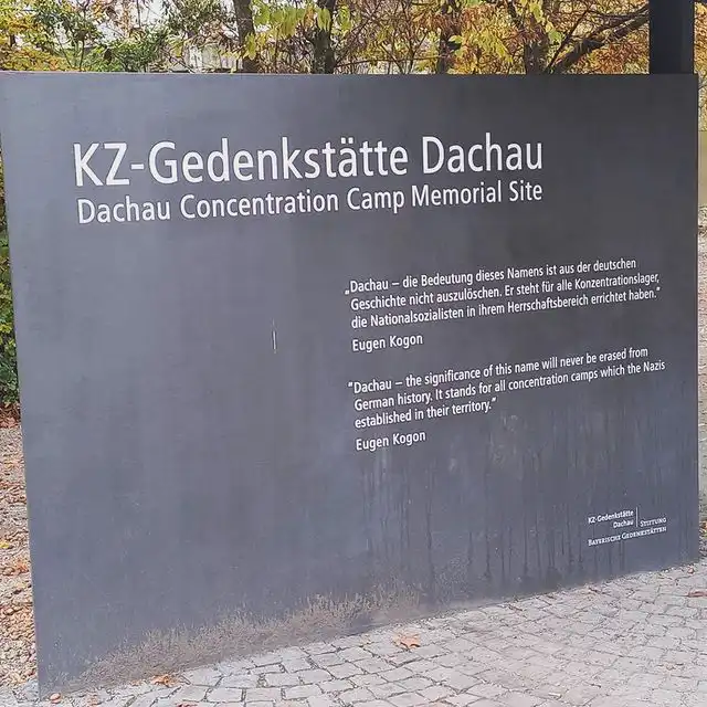 Gedenktafel von Dachau. In grauen Stahl ist folgende weiße Schrift eingelassen: KZ-Gedenkstätte Dachau. Dachau Concentration Camp Memorial Site. „Dachau – die Bedeutung dieses Namens ist aus der deutschen GEschichte nicht auszulöschen. Er steht für alle Konzentrationslager, die Nationalsozialsten in ihrenm Herrschaftsbereich errichtet haben. “ Eugen Kogon. “Dachau – the significance of this name will never be erased from German history. It stands for all concentration camps which the Nazis established in their territory.“ Eugen Kogon Unten rechts ist das Logo der Gedenkstätte eingelassen. Die Tafel ist bildfüllend. Vor der Tafel ist der gepflasterte Boden erkennbar. Im Hintergrund stehen Bäume