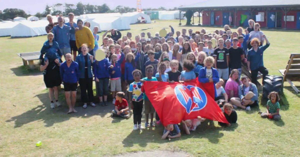 Gruppenbild auf dem Zeltlagerplatz „Unsere Welt Föhr“. Auf einer gelben Wiese mit zahlreichen Grünen Flecken stehen ca. 100 Personen, großteils Kinder, in Blauhemden vor einer großen Falkenfahne. Im Hintergrund sind weiße Großzelte und ein Gebäude zu sehen.