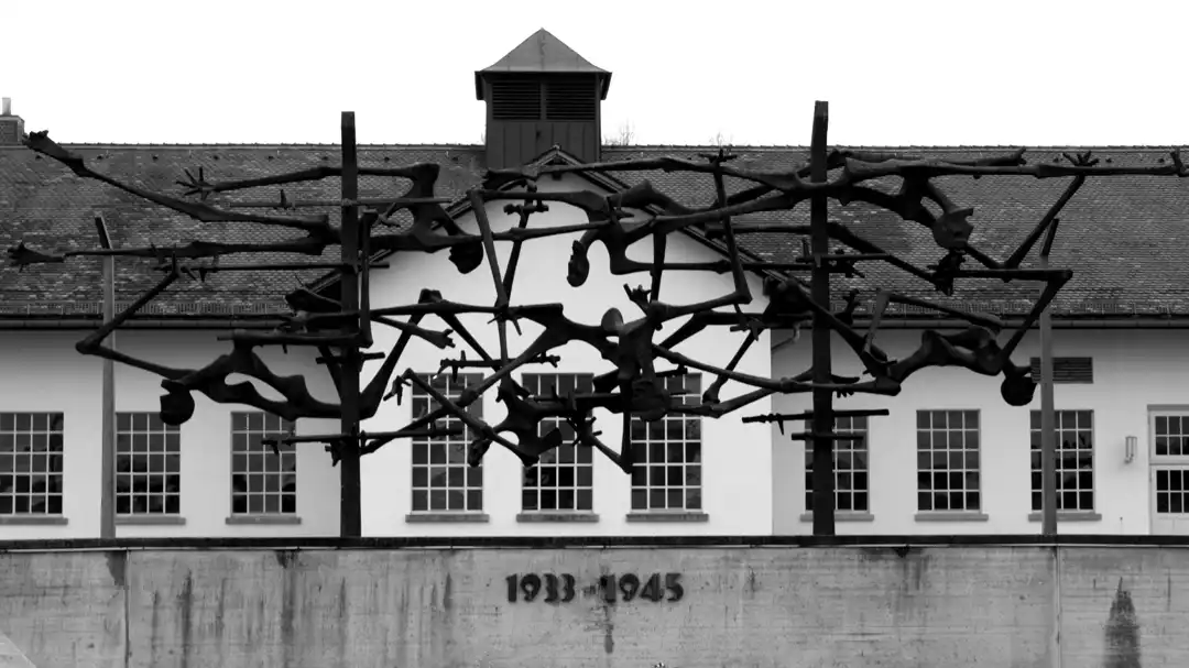 Schwarzweißphoto des internationalen Mahnmals „Menschen in Stacheldraht“ von Nandor Glid. Im Hintergrund das Hauptgebäude des KZ Dachau